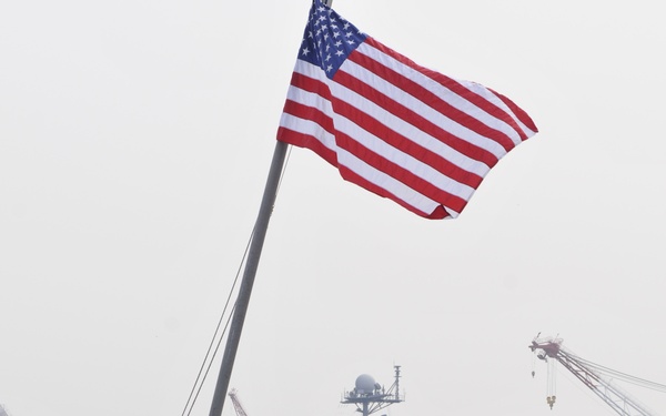 A U.S. Navy scene at a harbor showing two sailors in blue uniforms and hard hats standing on a smaller vessel with an American flag flying from a mast. In the background, a large aircraft carrier bearing the hull number '73' is moored at the pier, identifiable as the USS George Washington (CVN-73). Industrial cranes and green hillside vegetation are visible in the background, suggesting this is likely Yokosuka Naval Base, Japan.