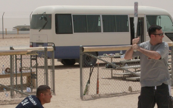 A baseball/softball game is being played on a sandy, desert-like field, likely at a military base given the arid environment and institutional setting. A batter holding an Easton bat stands at home plate while a catcher crouches behind, wearing a mitt. Several spectators watch from behind a chain-link fence near bleachers, with a white passenger bus visible in the background.