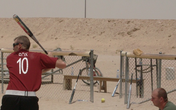A softball game is being played on a sandy, desert terrain, likely at a military base given the arid environment and makeshift field. A batter wearing a red jersey numbered 10 with 'SOX' on the back is in a batting stance, while a catcher crouches behind home plate wearing a mitt. Several other individuals are visible in the background near a dugout area.