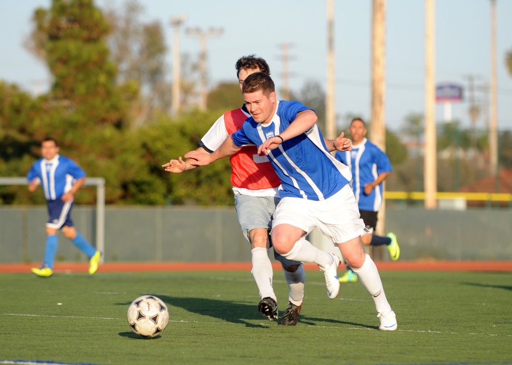 A competitive soccer match is taking place on an artificial turf field. Two players in the foreground are engaged in a physical challenge for the ball, with one wearing a blue jersey and white shorts and the other wearing a red training bib over a white shirt. Additional players and a goal post are visible in the background.
