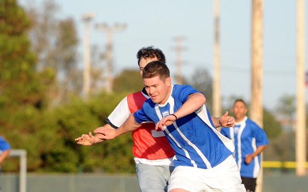 A competitive soccer match is taking place on an artificial turf field. Two players in the foreground are engaged in a physical challenge for the ball, with one wearing a blue jersey and white shorts and the other wearing a red training bib over a white shirt. Additional players and a goal post are visible in the background.