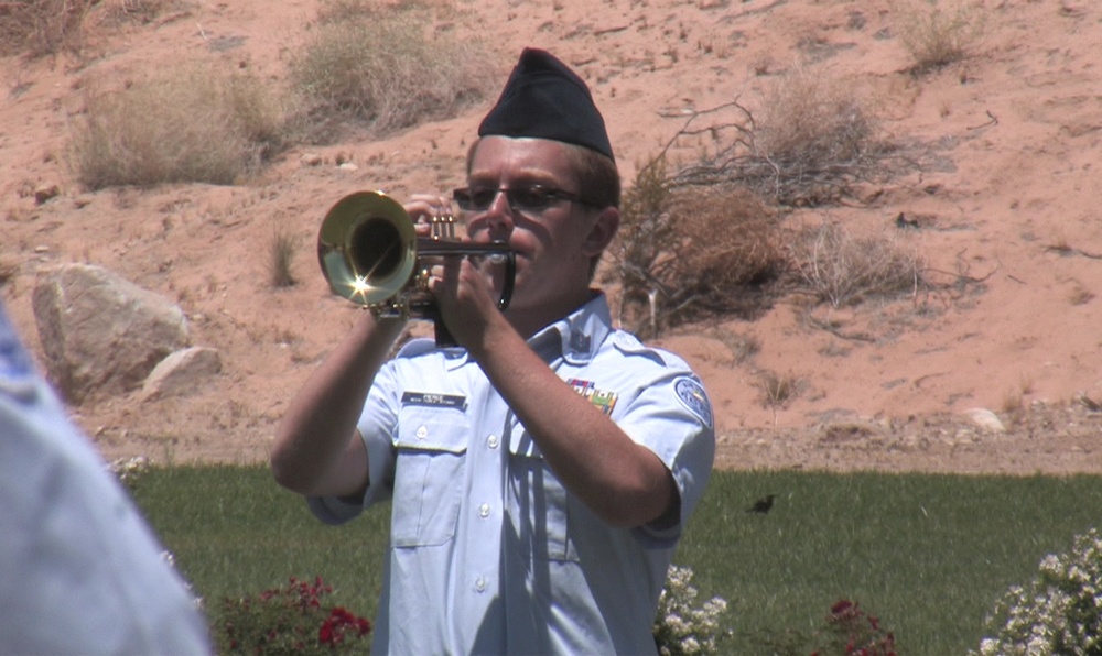A military service member in a light blue uniform is playing a brass bugle or trumpet, likely performing 'Taps' or a ceremonial call at what appears to be an outdoor memorial or funeral service. The setting features a desert landscape with red rock formations in the background and a manicured lawn with flowers in the foreground. The occasion appears to be a formal military ceremony.