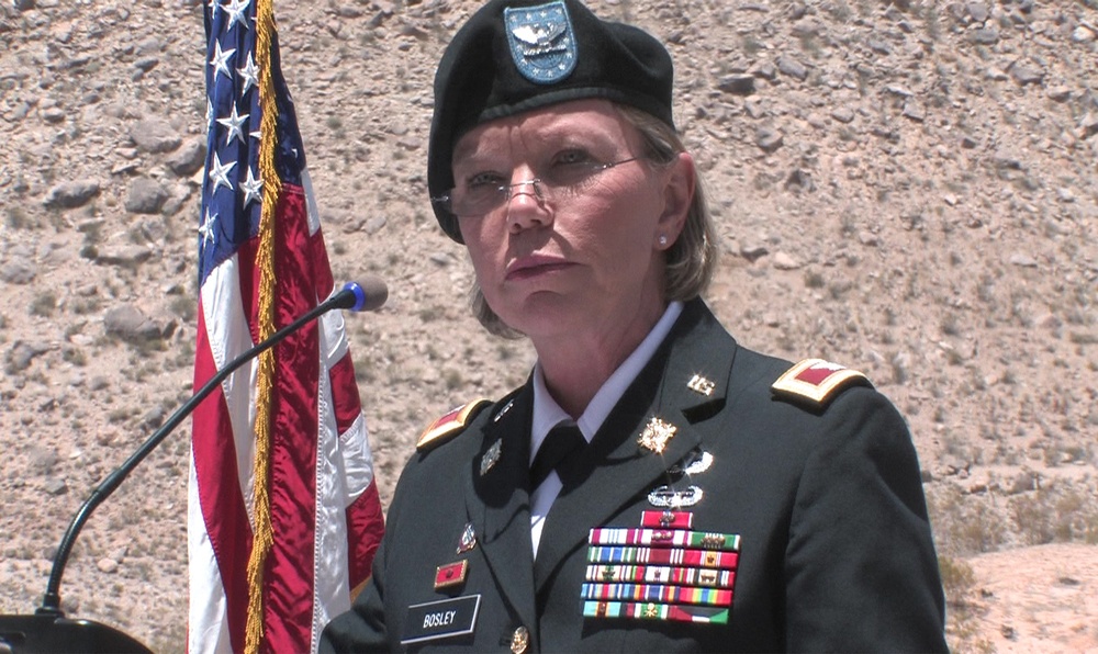 A U.S. Army officer identified by a name tag reading 'BOSLEY' stands at a podium with a microphone, wearing a dress green uniform with multiple service ribbons and medals. She is wearing a black beret with a unit flash and rank insignia visible on her shoulders. An American flag with gold fringe is positioned to her left against a rocky outdoor backdrop.