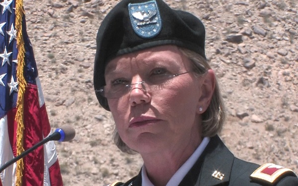 A U.S. Army officer identified by a name tag reading 'BOSLEY' stands at a podium with a microphone, wearing a dress green uniform with multiple service ribbons and medals. She is wearing a black beret with a unit flash and rank insignia visible on her shoulders. An American flag with gold fringe is positioned to her left against a rocky outdoor backdrop.