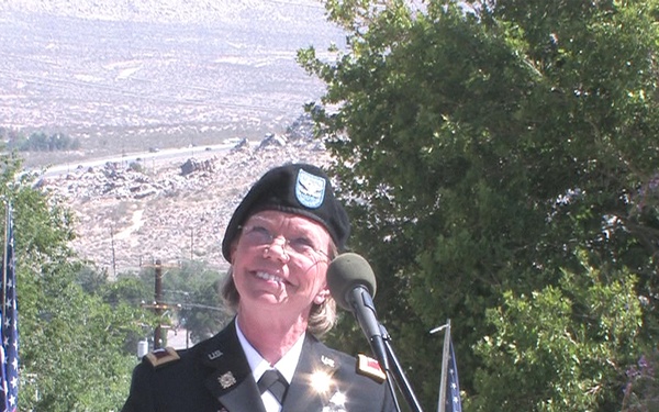 A female U.S. Army officer in dress uniform is speaking at a podium during what appears to be an outdoor military or patriotic ceremony. She is wearing a black beret and dress blues with multiple service ribbons and medals. American flags are visible on both sides of the podium, and the setting appears to be an elevated outdoor venue with a desert landscape in the background.