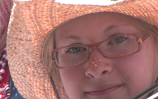 A young woman wearing a straw cowboy hat and red-framed glasses is playing a violin or fiddle. She is dressed in patriotic American-themed attire, including a red, white, and blue beaded necklace and a stars-and-stripes patterned garment. The close-up shot captures her focused expression during what appears to be an outdoor performance.