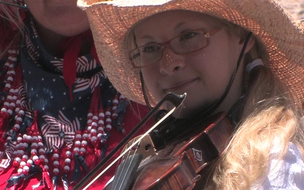 A young girl wearing a straw hat and glasses is playing a violin outdoors. She is dressed in a white shirt and appears to be performing at an outdoor event, likely in an arid or desert setting. An adult in a red sweater with patriotic red, white, and blue beaded necklace is seated behind her.