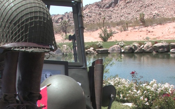 The image shows a display of World War II-era U.S. military equipment and gear arranged on what appears to be a military vehicle, likely at an outdoor event or exhibition. The setting includes a desert landscape with rocky hills, a small pond or water feature, and flowering shrubs in the background. The collection includes helmets, boots, pouches, and other period-authentic military accessories.