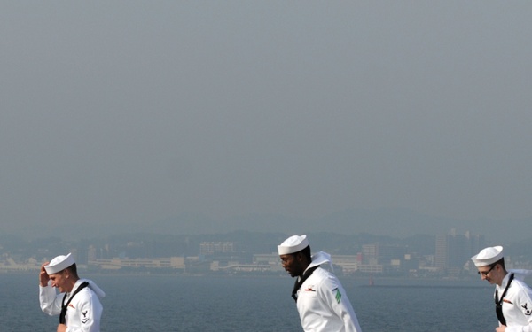 Five U.S. Navy sailors dressed in white dress uniforms are walking across what appears to be the flight deck of an aircraft carrier. Several sailors are adjusting their white dixie cup covers (hats), likely due to wind. A coastal city or naval base is visible in the hazy background across the water.