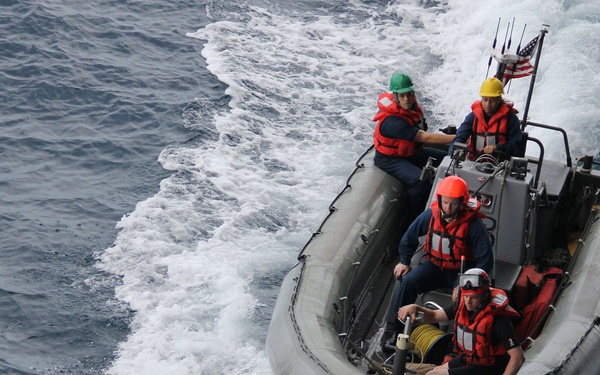 A rigid inflatable boat (RIB) is underway at speed on open ocean water, carrying five personnel wearing orange life vests and colored safety helmets. The boat appears to be military or coast guard in nature, indicated by the U.S. flag visible at the stern and the gray hull construction. The vessel is creating a significant wake, suggesting high-speed travel.