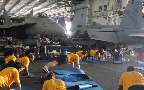 A group of military personnel in yellow t-shirts and dark shorts are performing push-ups and exercise routines on blue yoga mats inside what appears to be an aircraft carrier hangar bay. Multiple fighter jets, including aircraft marked '405' and '503', are visible in the background. The scene depicts a physical training (PT) session conducted in the ship's hangar deck.