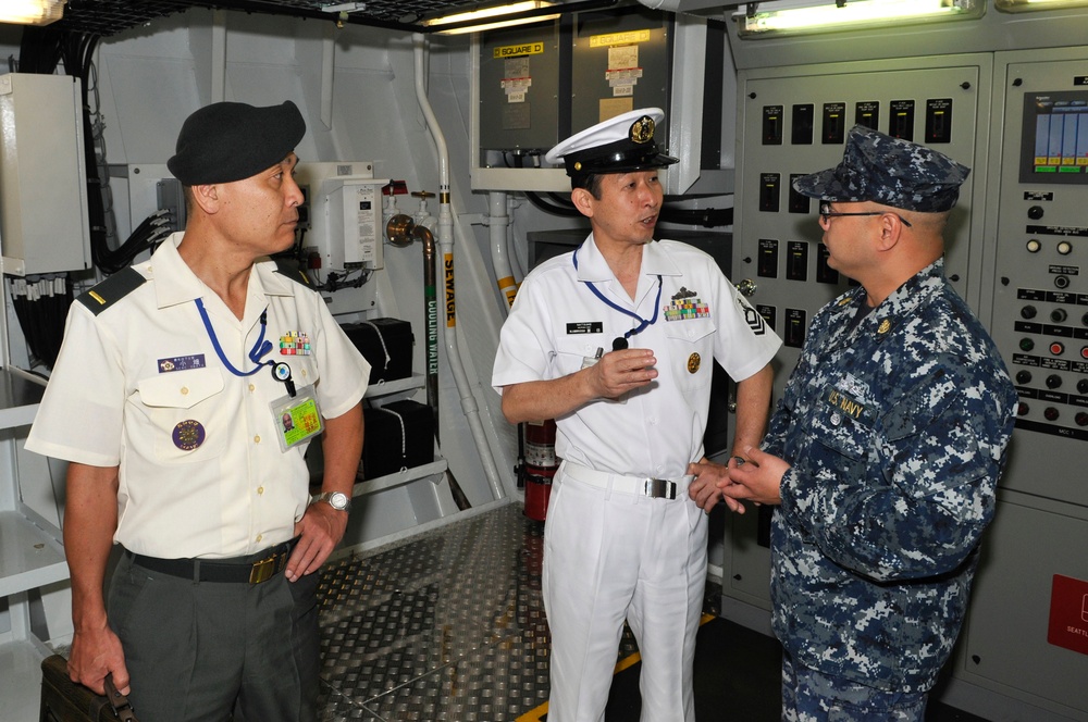 Three military personnel are engaged in conversation inside what appears to be a naval vessel's mechanical or engineering compartment. Two individuals are wearing white naval uniforms with insignia consistent with Asian naval forces, while the third wears U.S. Navy blue digital camouflage (NWU Type I) utilities. The setting includes control panels, piping labeled 'SEWAGE' and 'COOLING WATER', and various shipboard equipment.
