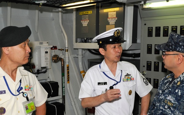 Three military personnel are engaged in conversation inside what appears to be a naval vessel's mechanical or engineering compartment. Two individuals are wearing white naval uniforms with insignia consistent with Asian naval forces, while the third wears U.S. Navy blue digital camouflage (NWU Type I) utilities. The setting includes control panels, piping labeled 'SEWAGE' and 'COOLING WATER', and various shipboard equipment.
