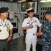 Three military personnel are engaged in conversation inside what appears to be a naval vessel's mechanical or engineering compartment. Two individuals are wearing white naval uniforms with insignia consistent with Asian naval forces, while the third wears U.S. Navy blue digital camouflage (NWU Type I) utilities. The setting includes control panels, piping labeled 'SEWAGE' and 'COOLING WATER', and various shipboard equipment.
