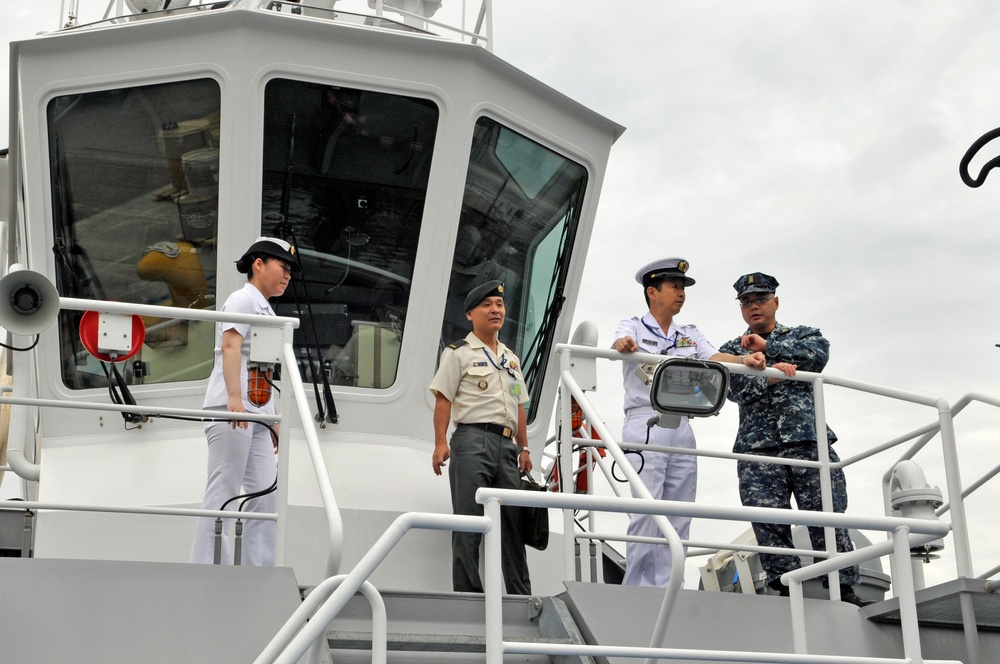Four military personnel are standing on the deck of a gray naval or coast guard vessel. Three individuals are wearing white naval uniforms with peaked caps, while one person is wearing a tan/khaki military uniform and another is in a blue digital camouflage uniform. They appear to be engaged in a briefing or tour of the vessel.