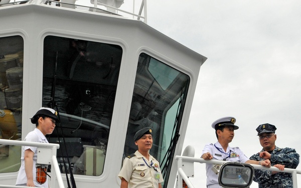 Four military personnel are standing on the deck of a gray naval or coast guard vessel. Three individuals are wearing white naval uniforms with peaked caps, while one person is wearing a tan/khaki military uniform and another is in a blue digital camouflage uniform. They appear to be engaged in a briefing or tour of the vessel.