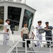 Four military personnel are standing on the deck of a gray naval or coast guard vessel. Three individuals are wearing white naval uniforms with peaked caps, while one person is wearing a tan/khaki military uniform and another is in a blue digital camouflage uniform. They appear to be engaged in a briefing or tour of the vessel.