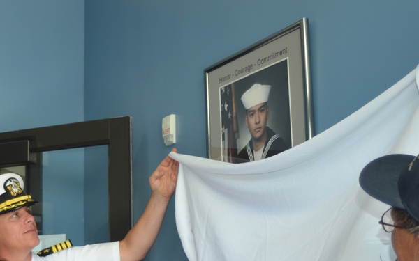 Three individuals are unveiling a framed portrait of a U.S. Navy sailor mounted on a blue wall. A naval officer in white dress uniform with rank insignia and medals participates alongside a woman in a red blouse and an older man in a baseball cap. The framed photo displays a young sailor in dress whites with an American flag background, beneath text reading 'Honor - Courage - Commitment'.