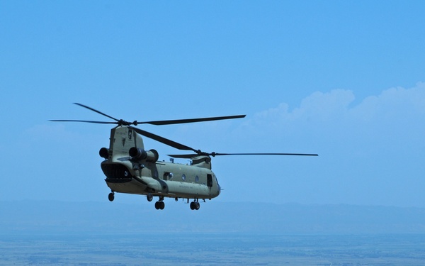 A military tandem-rotor helicopter, identifiable as a Boeing CH-47 Chinook, is captured in flight over a vast arid, semi-desert landscape with rolling hills and dry riverbeds visible below. The helicopter is painted in olive drab/tan military camouflage consistent with U.S. Army livery. The terrain and environmental conditions suggest an operational environment consistent with Central Asia or the Middle East.