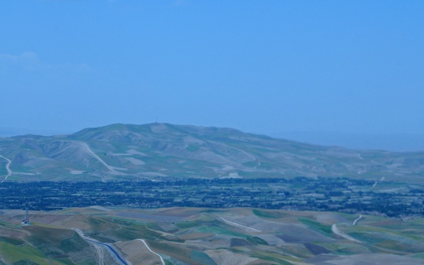 An aerial or elevated view of a rolling, semi-arid landscape featuring a prominent water canal or aqueduct cutting through undulating hills and agricultural fields. A valley with what appears to be a town or settlement is visible in the middle distance, backed by a large rounded mountain or hill. The image has a blue-tinted haze, suggesting atmospheric conditions or a blue color cast from the camera.