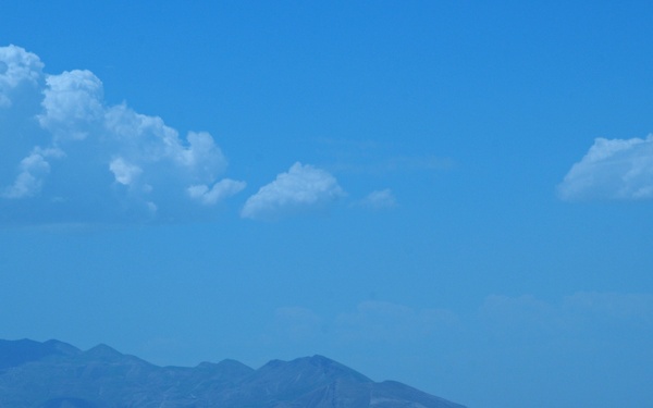 A wide-angle landscape photograph showing rolling arid hills in the foreground with a mountain range in the middle distance. The terrain appears dry and semi-arid with sparse vegetation, and a small settlement is faintly visible at the base of the mountains. The sky is bright blue with scattered cumulus clouds.