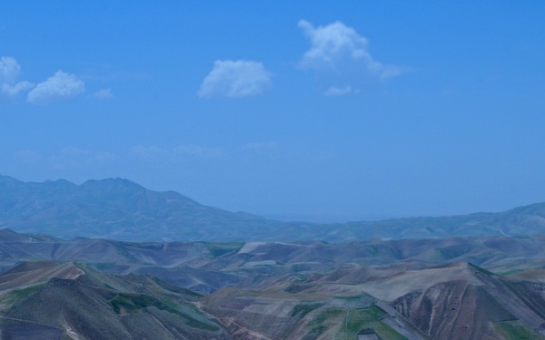 An aerial or elevated view of a vast, semi-arid mountainous landscape with terraced hillsides and winding dirt roads. The terrain shows a patchwork of cultivated fields and bare earth across rolling hills and ridgelines. A higher mountain range is visible in the hazy background under a partly cloudy blue sky.