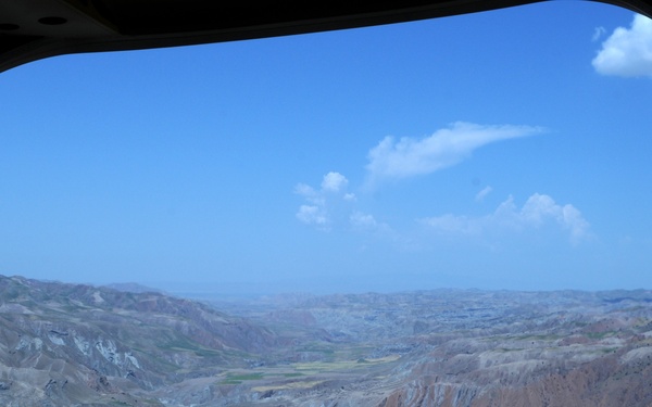 The image is taken from inside a military aircraft, looking out through an open rear cargo ramp door at a rugged, arid mountainous landscape below. A small settlement or village is visible nestled in a valley between eroded ridgelines with sparse green patches. The terrain is characteristic of Central Asian or Middle Eastern highland regions.