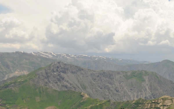 An aerial or high-elevation view of a rugged mountain landscape with multiple ridgelines and valleys. The terrain shows a mix of green vegetation, exposed rocky slopes, and terraced agricultural fields carved into the hillsides. Snow patches are visible on distant higher peaks, and large cumulus clouds dominate the upper sky.