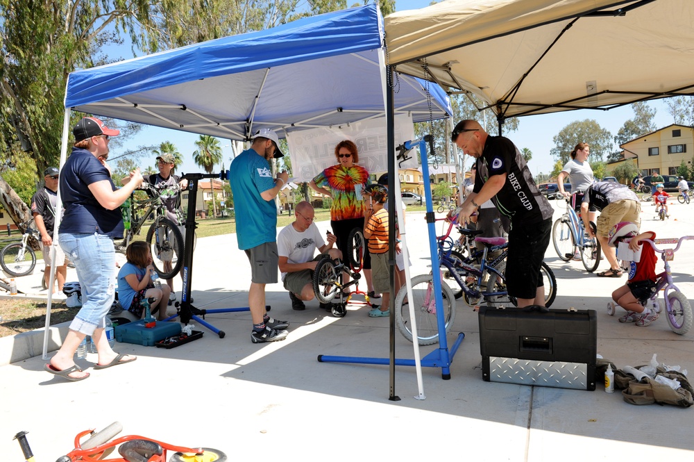 An outdoor community bike repair and maintenance event taking place under pop-up canopy tents in what appears to be a park or recreational area. Multiple volunteers and participants are working on bicycles of various sizes, including children's bikes. The event appears organized, with tool boxes, bike repair stands, and informational materials visible.