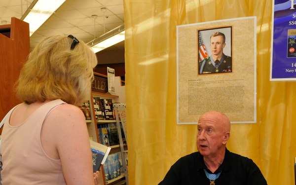 A book signing event is taking place at the Navy Exchange Yokosuka Book Store on May 19th, 2012, featuring Medal of Honor recipients SGT Don J. Jenkins and SSGT Kenneth E. Stumpf. An older bald man wearing a black polo shirt with a Medal of Honor around his neck sits at a signing table speaking with a woman. The event promotes the book 'Medal of Honor: Portraits of Valor Beyond the Call of Duty.'