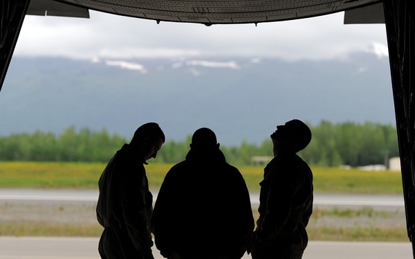 Three individuals are silhouetted against the open rear cargo ramp of a military transport aircraft, likely a C-17 or C-130, parked on a flight line. The background reveals a runway, green treeline, and snow-capped mountains, suggesting a northern or Alaskan airfield setting. The figures appear to be in conversation, with one looking upward toward the aircraft structure.