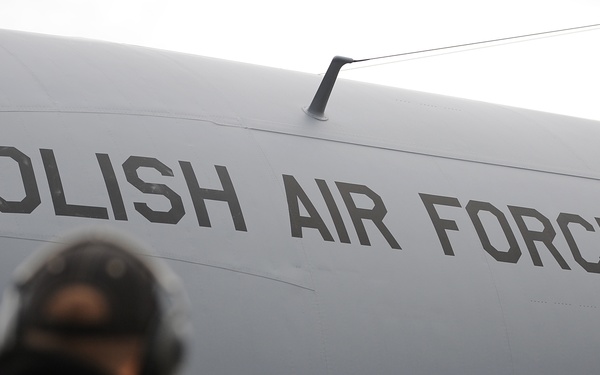 The image shows a large gray military transport aircraft belonging to the Polish Air Force, with 'POLISH AIR FORCE' prominently stenciled on its fuselage. A uniformed personnel member, visible from behind and slightly out of focus, stands in the foreground facing the aircraft. An open door/hatch with yellow interior framing is visible on the aircraft's side.