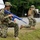 Two military personnel in U.S. Navy Working Uniform (NWU) camouflage are conducting a tactical training exercise in an outdoor gravel and grass area. The foreground figure is kneeling beside a white vehicle holding a blue training rifle (inert/dummy weapon used for drills), while a second figure is moving in the background. The bright blue color of the firearm confirms this is a non-live training scenario using a simulated weapon.