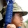 A female soldier in digital camouflage uniform and boonie hat holds a blue training/dummy rifle at the ready position. The image is a close-up profile shot emphasizing her focused expression and military equipment. The blue-colored weapon indicates this is a training or drill scenario using a non-live replica firearm.