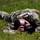 A person wearing military camouflage uniform is lying prone on green grass while operating a Nikon DSLR camera. The individual appears to be a military photographer capturing images from a ground-level perspective. Sunglasses are visible on their face, and the setting appears to be an outdoor grassy area in daylight.