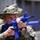 A military personnel wearing US Navy digital camouflage (NWU Type III) and a boonie hat is aiming a blue training/dummy rifle during what appears to be a tactical training exercise. The bright blue color of the weapon indicates it is a non-functional training replica, commonly used in military drills to distinguish training equipment from live weapons. A second similarly dressed individual is visible in the blurred background.
