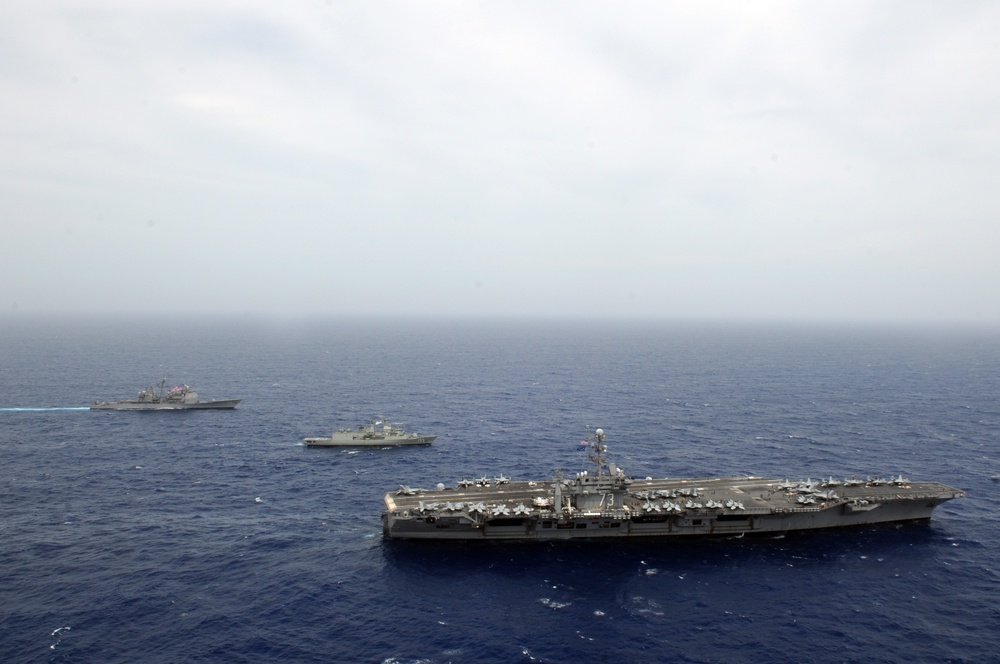 An aerial photograph showing a naval formation of at least three warships underway in open blue ocean under overcast skies. The foreground features a large aircraft carrier bearing the hull number '73', identifiable as USS George Washington (CVN-73), with numerous aircraft visible on its flight deck. Two additional escort vessels — likely a cruiser or destroyer and a supply/amphibious ship — are visible in the middle and background.