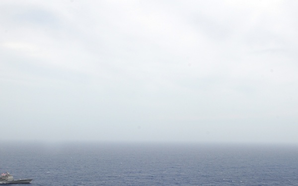 An aerial photograph showing a naval formation of at least three warships underway in open blue ocean under overcast skies. The foreground features a large aircraft carrier bearing the hull number '73', identifiable as USS George Washington (CVN-73), with numerous aircraft visible on its flight deck. Two additional escort vessels — likely a cruiser or destroyer and a supply/amphibious ship — are visible in the middle and background.