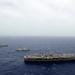 An aerial photograph showing a naval formation of at least three warships underway in open blue ocean under overcast skies. The foreground features a large aircraft carrier bearing the hull number '73', identifiable as USS George Washington (CVN-73), with numerous aircraft visible on its flight deck. Two additional escort vessels — likely a cruiser or destroyer and a supply/amphibious ship — are visible in the middle and background.