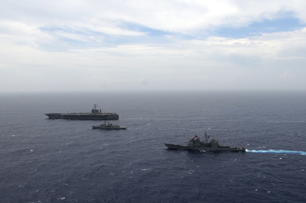 An aerial photograph shows three naval vessels underway in open ocean, likely during a formation exercise or transit operation. The formation includes a large aircraft carrier in the background, a smaller warship in the middle distance, and what appears to be a guided-missile cruiser or destroyer in the foreground. The ships are sailing in a loose column or echelon formation under partly cloudy skies.
