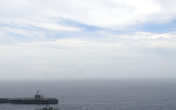 An aerial photograph shows three naval vessels underway in open ocean, likely during a formation exercise or transit operation. The formation includes a large aircraft carrier in the background, a smaller warship in the middle distance, and what appears to be a guided-missile cruiser or destroyer in the foreground. The ships are sailing in a loose column or echelon formation under partly cloudy skies.