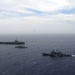 An aerial photograph shows three naval vessels underway in open ocean, likely during a formation exercise or transit operation. The formation includes a large aircraft carrier in the background, a smaller warship in the middle distance, and what appears to be a guided-missile cruiser or destroyer in the foreground. The ships are sailing in a loose column or echelon formation under partly cloudy skies.