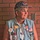 An older woman stands against a brick wall wearing a denim vest covered in patriotic pins, patches, and badges. She wears a stars-and-stripes baseball cap and displays numerous military-related insignia. Her vest identifies her as 'Kay Erickson' nicknamed 'Sam' affiliated with the Patriot Guard NW Metro.