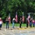 A group of people are gathered outside the Roseville National Guard Armory, holding multiple American flags in what appears to be a patriotic ceremony or event. The participants are lined up along a parking lot in front of the brick building, with many holding flag poles displaying U.S. flags. The setting suggests a formal or commemorative gathering, possibly honoring veterans or military personnel.