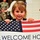 A young girl, approximately 3-4 years old, holds a welcome home sign for someone named Justin. The sign features an American flag graphic and reads 'Welcome Ho[me] JUSTIN - We Missed Yo[u]'. The setting appears to be a military homecoming event, suggested by a person in camouflage uniform visible in the background.
