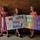 Three children stand against a brick wall holding handmade welcome home signs for their father. The left child holds a small American flag, suggesting a military homecoming context. The signs read 'WE LOVE YOU DAD!' and 'WELCOME HOME DAD!' indicating a reunion celebration.