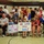 A large group of people, including many children, are gathered in what appears to be a gymnasium or community center for a military homecoming welcome event. Two handmade signs are prominently displayed: one reads 'WELCOME HOME SANGLE JOHN' and the other reads 'WE LOVE YOU DAD!' in colorful letters. The atmosphere appears celebratory and emotional, consistent with a military return or deployment homecoming ceremony.