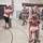 A military homecoming ceremony taking place inside what appears to be a gymnasium or armory. Multiple soldiers in U.S. Army camouflage (OCP/MultiCam) uniforms are being reunited with family members, including emotional embraces with children and adults. American flags are displayed in the background, and onlookers including a person in a wheelchair are visibly emotional.