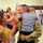 A U.S. Army soldier in camouflage uniform is being reunited with his family, holding a child in his arms while a woman smiles joyfully beside him. The setting appears to be an indoor military homecoming ceremony, with American flags visible in the background. A video camera operator is capturing the emotional reunion.