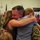 A military service member in camouflage uniform is embracing two children in an emotional reunion, likely following a deployment. The scene takes place inside what appears to be a military facility or gymnasium decorated with American flags. Other soldiers and civilians are visible in the background, suggesting a larger welcome-home ceremony.